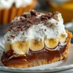 A slice of Banoffee Pie on a white plate showing layers of caramel, banana slices, whipped cream, and chocolate shavings on a biscuit crust.