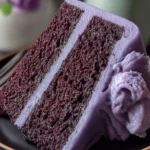 Slice of purple cake with lavender frosting on a black plate next to a white ceramic dinosaur.